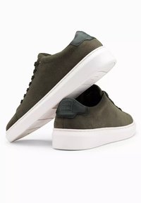 Baskets en suede vert olive avec semelles en caoutchouc blanches, dotées de lacets ronds et d'une languette de talon vert foncé avec des accents de marque.