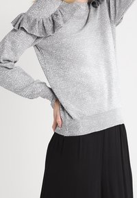 Grauer Pullover mit gerafftem Kragen und langen Ärmeln, mit einem gerippten Saum, getragen über einem schwarzen Rock. Subtil strukturiertes Material.