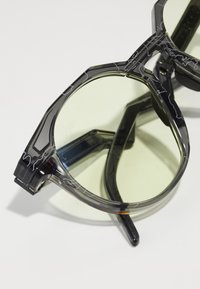 Sonnenbrille mit einem geometrischen schwarzen Rahmen, der ein klares Muster aufweist, grünen Gläsern und dicken Bügeln. Das Design enthält filigrane Linienverzierungen.