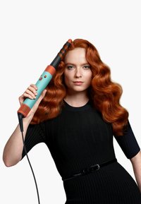 Hårkrullningsstång med en turkos och orange design, konform, med texturerat grepp och en lång strömkabel, hålls av en person med vågigt rött hår.
