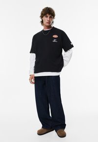 Ung mand med krøllet hår iført en lagdelt sort og hvid langærmet skjorte, løse mørke jeans, brune slip-on sko og en sølv kædedyse.