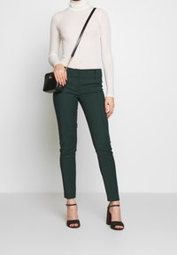 Figurpassande gröna byxor tillsammans med en ljus, långärmad polotröja. Accessoarer inkluderar en liten svart axelväska och svarta klackar.