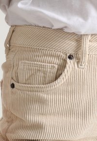 Beige corduroy pants with vertical ribbing, featuring a curved back pocket and black metal button. Smooth texture with a fitted design.
