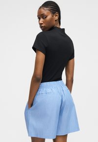 Γυναίκα με κοτσίδες, φορώντας μαύρο κοντομάνικο τοπ και μπλε ψηλόμεσο ριγωτό σορτς με τα χέρια στις τσέπες, που κοιτά ζόρι.