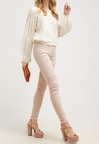 Krämfärgad blus med spetsdetaljer, ljusrosa tajta jeans och sandfärgade plattformsskor. Modellen håller en brun clutch och visar upp en avslappnad outfit.