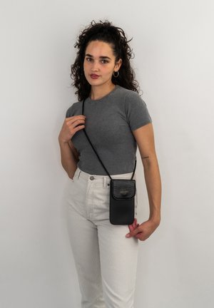Schwarze Umhängetasche mit Krokodilprägung auf dem Deckel, aus Leder gefertigt; getragen mit einem langen Gürtel von einem Modell in einem grauen Oberteil und weißen Hosen.