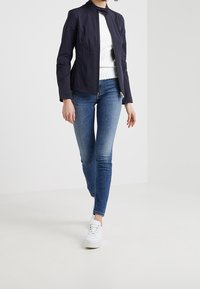 Navy jacket with a fitted design, high collar, and zip closure; paired with a white ribbed shirt and blue skinny jeans. White sneakers.