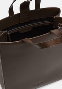 Brązowa torba typu tote z gładką fakturą, podwójnymi uchwytami u góry i zamykaną na zamek kieszenią wewnętrzną. Na boku znajduje się wytłoczone logo.