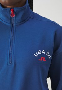 Sudadera de forro polar azul con un cuello alto, que presenta una cremallera roja y un logo "USA 24" en blanco en el pecho con un acento rojo debajo.
