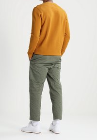 Suéter mostaza de manga larga combinado con pantalones verde oliva, con un corte relajado. Lleva puesta unas zapatillas blancas y calcetines hasta los tobillos.