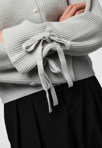 Hele halli kudumüts ribitekstuuriga, millel on mansetil side. Mustad volditud püksid on osaliselt nähtavad alt.