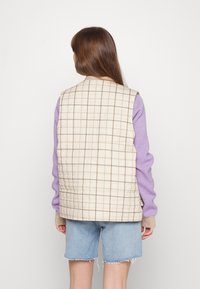 Vadderad beige väst med brun rutad mönster, lila långärmade tröja, ljusblå denimshorts och en påtaglig yta.