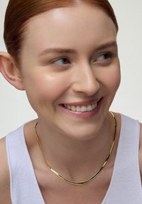 Złota metalowa naszyjnik o gładkim, zakrzywionym kształcie z delikatnym skrętem. Noszony na szyi, na górnej części bez rękawów w kolorze białym. Gładkie wykończenie.