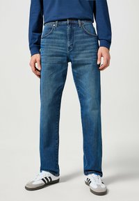 Niebieskie dżinsy z prostym krojem nogawki, pięcioma kieszeniami i widocznymi szwami. Połączone z białymi i szarymi sneakersami.