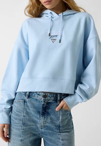Sweat à capuche court bleu clair avec cordon de serrage, présentant le logo brodé "Guess Jeans" et un patch triangulaire. Associé à un jean en denim délavé.