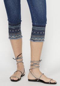 Capris en denim avec des poignets embellis en tissu bleu foncé, présentant des motifs complexes et des ourlets effilochés. Sandales plates noires avec des brides entrecroisées.