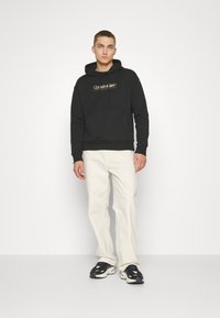 Felpa nera con tasca frontale e logo dorato, abbinata a pantaloni larghi di colore chiaro e sneaker nere, che presentano un outfit casual.