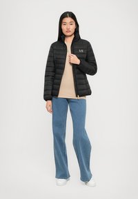 Zwarte gewatteerde jas met een ritssluiting aan de voorkant, een hoge kraag en een logoprint op de borst, gedragen over een beige top en blauwe flared jeans. Witte sneakers.