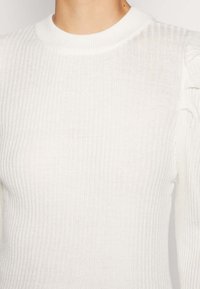 Pull en maille côtelée blanche avec un col rond et une texture subtile, silhouette ajustée et détails froncés aux épaules.
