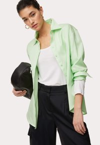 Lichtgroen oversized shirt met knopen, gedragen over een witte tanktop, in combinatie met zwarte broek en een zwart leren clutch.