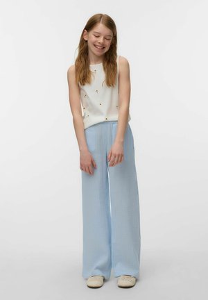Young girl with long hair wearing white sleeveless top with small flower prints, light blue wide pants, and white slip-on shoes, smiling.