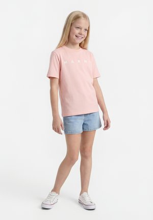 Smilende ung pige står iført en lyserød kortærmet skjorte, lyseblå denimshorts og hvide sneakers mod en hvid baggrund.