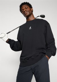 Melnās kokvilnas pelēkā jaka ar ribotiem piedurknēm un apakšmalu, uz krūšu ir neliels balts logotips. Modelis tur golfa nūju pāri pleciem.