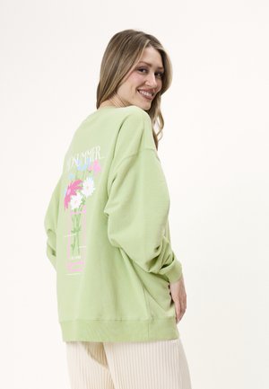 Kvinne smiler over skulderen iført en lys grønn oversized sweatshirt med et fargerikt blomstertrykk og tekst på baksiden.