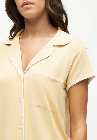Femme portant une chemise à manches courtes jaune clair avec des bordures blanches au col, à la poche et aux bords des manches.