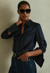 Camisa de satén azul marino con un gran cuello, botones en la parte delantera y mangas anchas, combinada con jeans de denim azul de talle alto y un cinturón negro.