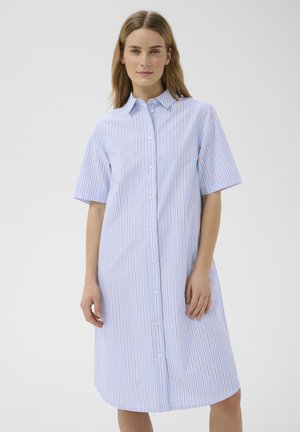 Femme portant une robe-chemise boutonnée à manches courtes et longueur genou avec des rayures verticales bleues et rouges, debout devant un fond uni.