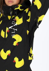 Zwarte jas met een herhaald patroon van gele eenden, voorzien van een ritssluiting aan de voorkant, verstelbare capuchon en trekkoorddetails.