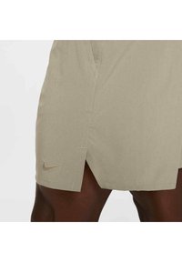 Pantalones cortos deportivos beige con una abertura lateral, que presentan un pequeño logo dorado de Nike en la parte inferior izquierda, hechos de una tela suave y ligera.