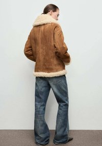 Giacca in suede marrone con colletto e orlo beige peloso, caratterizzata da una vestibilità rilassata, abbinata a jeans a gamba larga azzurri.