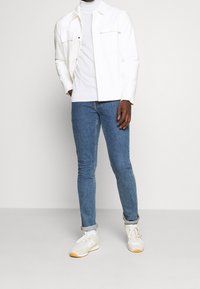 Uomo che indossa una giacca bianca e un dolcevita, pantaloni jeans blu e sneakers bianche, in piedi davanti a uno sfondo luminoso e semplice.