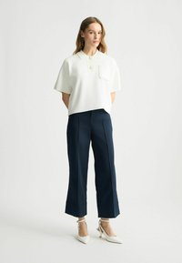 Bílý zkrácený polo top s krátkými rukávy a náprsní kapsou, kombinovaný s námořnickými kalhotami se širokými nohavicemi a bílými lodičkami se špičatou špičkou.