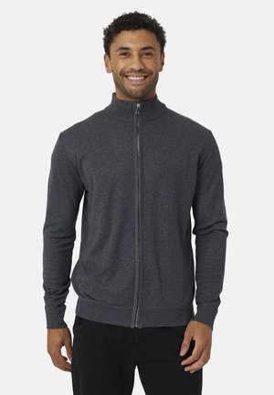 Uomo sorridente che indossa un maglione grigio scuro con zip integrale, polsini e orlo a coste, in piedi davanti a uno sfondo chiaro uniforme.