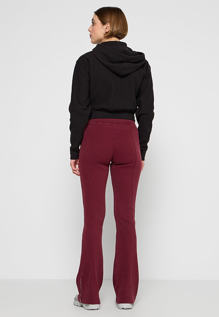 Persoon met de rug naar ons gekeerd, gekleed in een zwarte hooded sweater, bordeauxrode flare pants en witte sneakers, staand tegen een effen witte achtergrond.