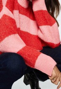 Kvinde iført en rød og pink stribet fuzzy sweater, mørke jeans og sorte ankelstøvler, med hånden hvilende nær knæet.