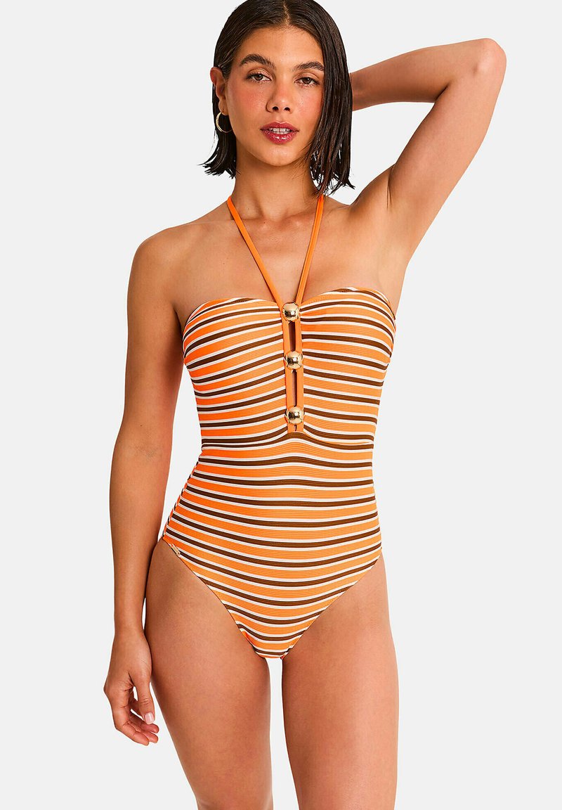 Femme portant un maillot de bain une pièce à rayures horizontales orange, blanc et marron, avec un col halter et des détails boutons dorés, posant avec un bras levé.