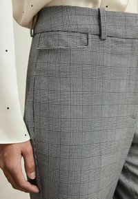 Prim-plan al unei persoane care poartă pantaloni cu talie înaltă, în carouri negre și albe, cu o mână de bluză cu buline vizibilă.
