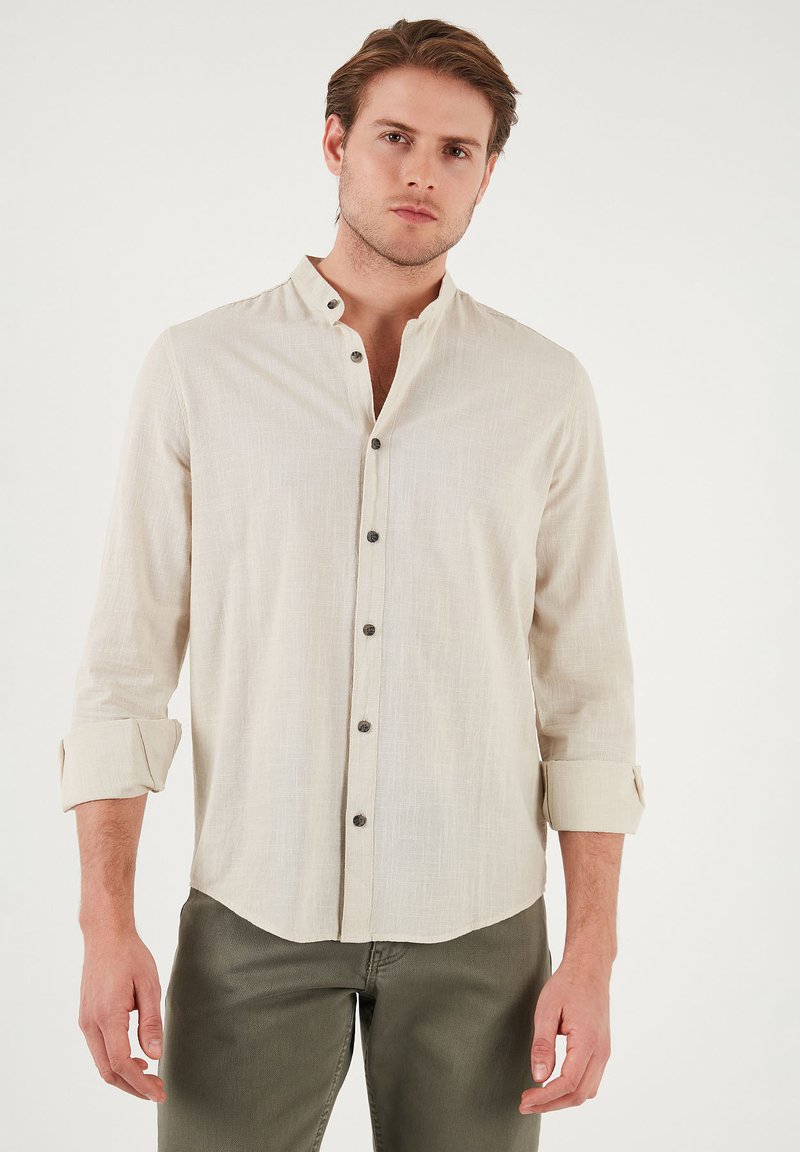 Chemise beige clair à manches longues avec un col montant, présentant des boutons marron sur le devant et des poignets retroussés, associée à un pantalon vert olive.