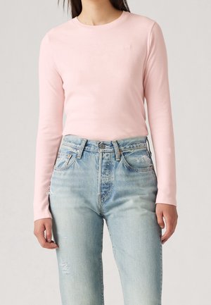 Person iført en tætsiddende, lyserød langærmet top og lyseblå, højttaljede, slidte jeans, stående foran en ensfarvet baggrund.