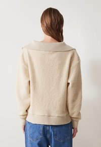 Maglione in pile beige texturizzato con un ampio colletto a coste, spalle scese e polsini elasticizzati. Indossato su jeans in denim blu a vita alta.