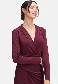 Femme aux cheveux bruns en chignon bas, portant une robe portefeuille à manches longues de couleur rouge profond, ornée de petits pois noirs, sur un fond uni.