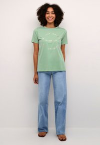 Groene katoenen T-shirt met korte mouwen, met witte tekst: "Geniet van elk moment van de zomer." Gecombineerd met lichtblauwe wijd-uitlopende jeans.