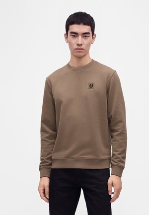 Ung mand iført en brun sweatshirt med et lille logo på brystet, parret med sorte bukser, stående mod en ensfarvet hvid baggrund.