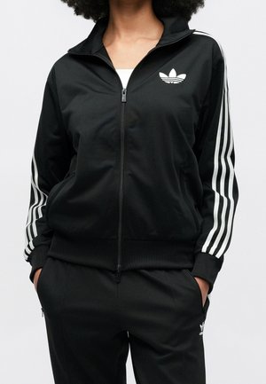 Person trägt einen schwarzen Adidas-Trainingsanzug mit weißen Streifen an Ärmeln und Hosen, Jacke mit Frontreißverschluss und Hände in den Taschen.