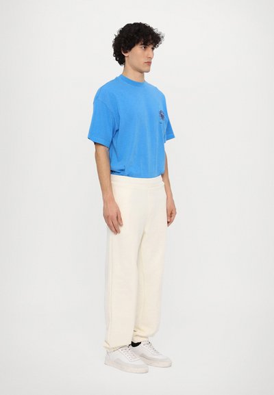 Blue cotton T-shirt with short sleeves, featuring a graphic print. Paired with white, soft-textured, tapered sweatpants and white sneakers.