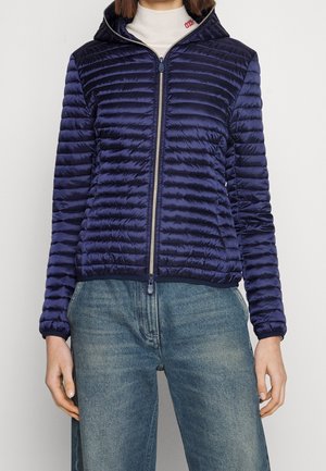 Femme portant une veste matelassée bleu marine zippée avec capuche, associée à un jean en denim bleu et un pull à col roulé blanc.